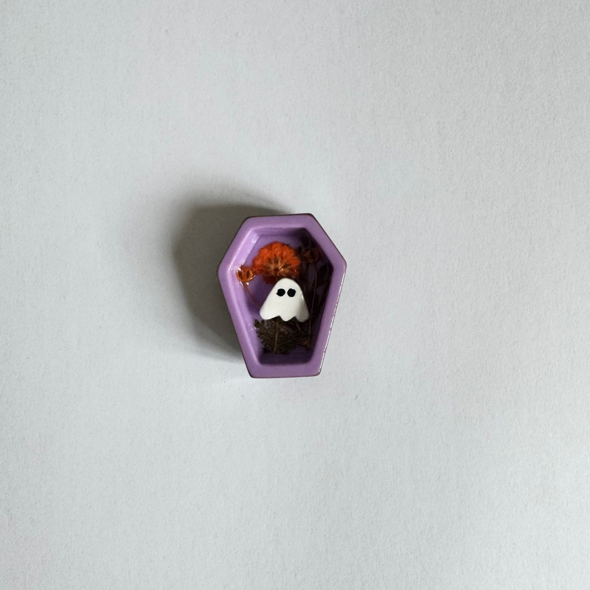 Purple coffin-shaped container with Halloween-themed items (dried flowers and clay sheet ghost) on a light gray background.